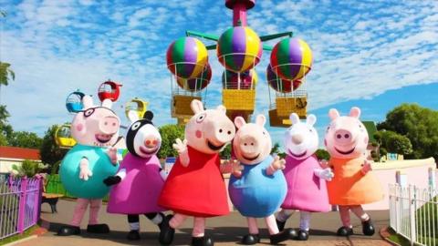 Line of Peppa Pig charachters in front of hot air balloon ride at theme park on a sunny day.