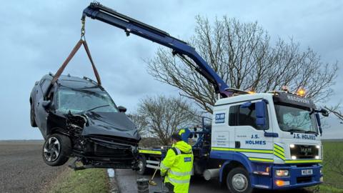 A van with a crane holding up a smashed up car. The van has neon stripes and "J.S. Holmes" and "recovery" on its front and side. The damaged car is black has a crunched front bonnet and heavily cracked window screen. They are on a road with fields and trees on either side. A man in a high-vis coat is facing towards the smashed car.