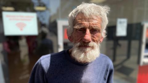 A man looks straight at the camera in a head and shoulders image. He is wearing a blue round-necked sweater and has grey hair and a beard. He is standing outside in full sun.