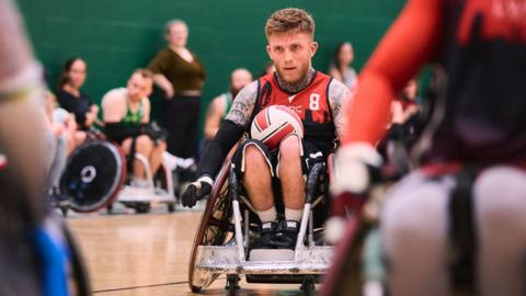 Joel Connor-Saunders covered in tattoos, wearing a red vest and playing wheelchair rugby. He is in front on two team mates and there are people out of focus watching in the background.