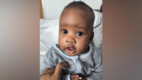 A young baby boy with short dark hair looks towards the camera. He is wearing a grey zip-up babygrow and is being held by an adult whose hand is visible.