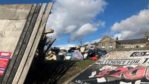 A broken fence with a big gap in the middle, revealing a mound of rubbish on some grass