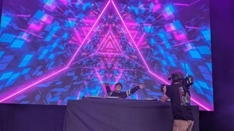 A stage showing lots of bright festival lights, with a cameraman taking footage of the DJ - who is smiling and has his hands out towards the audience 