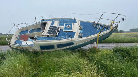 A cabin cruiser boat, about 6m (20ft) long and 2m (6ft) wide, lies on its side next to farmland, a narrow road can be seen in the background and more fields and trees beyond it.
