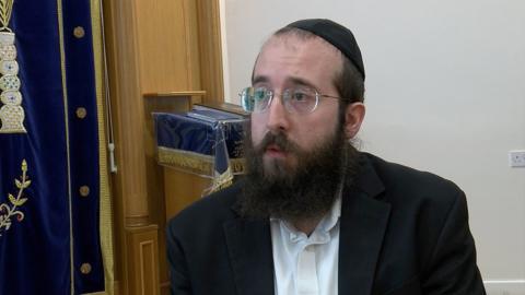 Rabbi Bentzion Alperowitz, looking left out of frame, wearing glasses and a yarmulke or traditional Jewish skullcap. He has a long beard and is wearing a black blazer and white shirt.