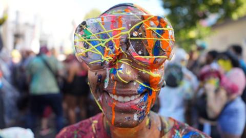 A person in clear goggles and a red t-shirt smiles down the lens of the camera, They are covered in colourful splatterings of paint. 