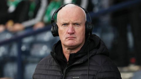 Kenny Macintyre in a football stadium wearing a black puffer jacket and black headphones