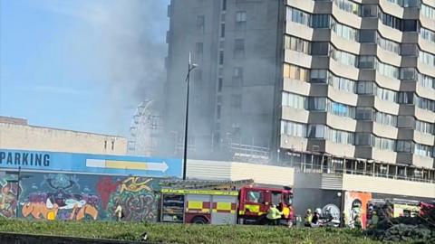 A tower block with some smoke outside the building. There are fire engines outside the building.