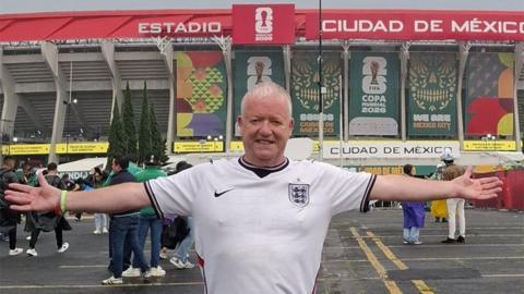 Paul Clegg in a white England shirt standing outside the Azteca Stadium in Mexico with his arms stretched out to the sides. He has short white hair 