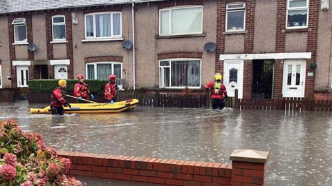 Emergency services staff take an inflatable boat to rescue people. The street is flooded with water which goes above the knees of rescue workers.