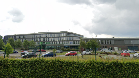 A Google streetview image of a school building which is set behind a green mesh fence and large green hedge. A number of cars are parked in front of the large grey building which features rectangular windows across three floors.