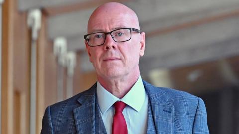 Kevin Stewart, a bald man with glasses, in the Scottish Parliament. He is wearing a grey suit, white shirt and red tie. 