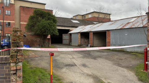 Police tape across the open gateway to a single-storey building with corrugated iron roof. Two large doors are open. Parked cars and other buildings can be sen beyond.
