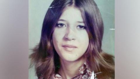 Laura Aimee, in a school photo from the 1970s. She has long brown hair parted down the middle and brown eyes, and wears a patterned blouse. 