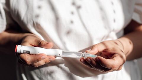 A woman wearing a white t-shirt holds a white injectable medicine pen in front of her stomach