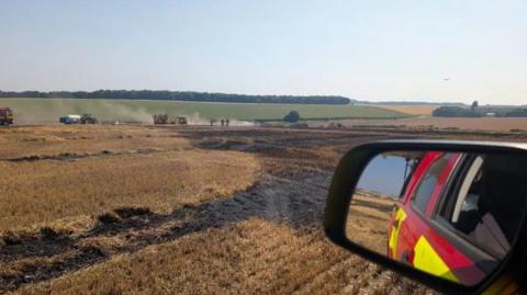 A view of a burned land from a fire rescue vehicle. Smoke is seen rising from the burnt ground. Three people are seen in the distance. It is a sunny day.