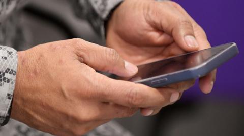 A person holding a mobile phone. The photo is a close-up of their hands and phone.