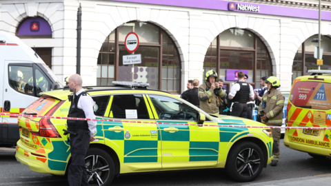 An ambulance car is parked outside a Natwest bank and in front is a red and white cordon. there is a police officer stood in front, and behind is a group of police officers and fire fighters.