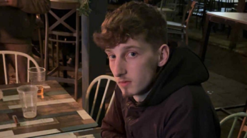 Mr Langley looks into the camera with an expression-less face. He is wearing a black hoodie and jacket, and is sat at a wooden table.