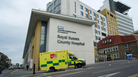 An ambulance passes a sign on the wall of a building that reads Royal Sussex County Hospital in Brighton. There are other vehicles to the left of the image. 