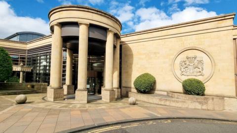 Exterior of High Court of Glasgow. A brown stone building with a round, pillared entrance and court of arms on the wall.