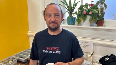 A man wearing a black T-shirt and holding a mug looks at the camera. Behind him is a window ledge with houseplants on, and to his right there is a yellow wall.