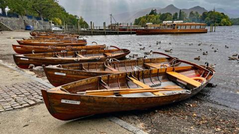 Derwentwater in Keswick. A line of wooden rowing boats are along the shore of the lake. Longer tourist boats are docked in the distance. A flock of ducks are swimming in the water.