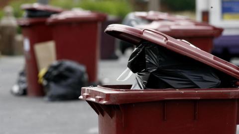 A close-up of a the lid of a red wheelie bin. The bin is full and the lid is partly open with a black bin bag visible in the top. In the background, there are several other red wheelie bins and bin bags on the floor around them.