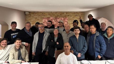 Nineteen men, of various ages, look jubilant as they pose for a photograph. One person, who is at the back and on the left-hand side, is making a 'V' for victory or peace sign.