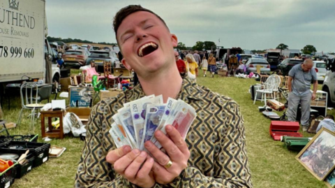 A young man stand at an outdoor sale. He holds a wad of cash in front of him. He is throwing back his head and laughing, with his eyes closed.