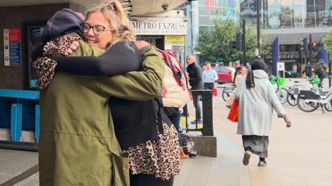 Mika Ap Ellis embraces her father outside a busy London Underground station entrance, surrounded by passersby and bicycles in the background.