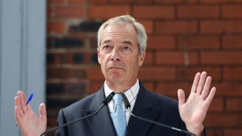 Reform UK leader Nigel Farage attends a press conference ahead of the Makerfield by-election, in Ashton-in-Makerfield in June. He wears a navy blue blazer, light blue tie and white shirt with blue and beige checks.