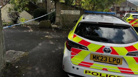 Police car, police tape and road sign which says Hillcroft North, Hillcroft South