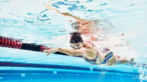 A woman wearing a swimming cap swimming in a swimming pool, seen from under the water level