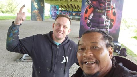 Nathan and Tony, both men, smiling for camera while taking a selfie in front of the artwork.
