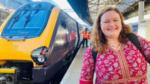 Woman - Aimee Smith - in bright maroon dress, smiling at camera, on rail platform, with train in the background.