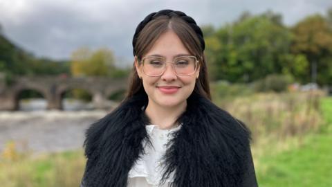 Molly is wearing a black padded alice band and glasses. She has long brown hair, centre parting, and is smiling whilst looking at the camera. She's wearing a furry black jacket and a white blouse with pearl buttons. She's standing in front of a river and grassy area. There's also an old stone bridge in the background. 