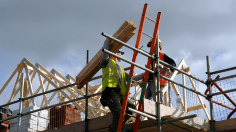 Two men in high visibility jackets and helmets on scaffolding with the wooden frame of a roof behind them
