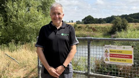A man with white hair is smiling. He is wearing a black t-shirt with a green logo that reads "environment agency". He is standing near metal fencing with warning signs on it. Beyond him is the river Severn surrounded by green and brown grass