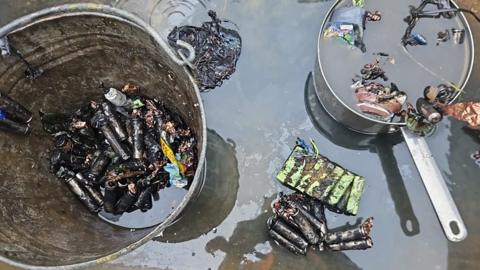 Aftermath of the fire with a metal bucket and saucepan filled with charred batteries and surrounded by water 
