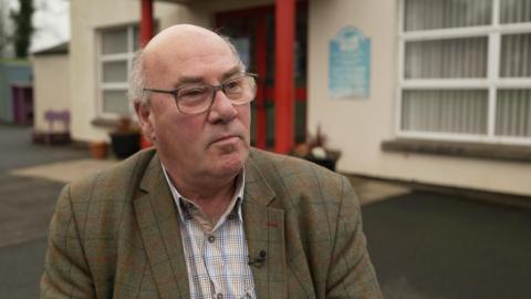 A man wearing a blue white and yellow shirt and a green blazer. He is looking to the right of the camera, and is wearing a pair of glasses. Behind him is a white building with a red porch. It could be a school.