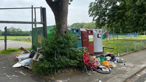 A row of large recycling bins in a public car park with an assortment of items, including plastic bags, boxes and a camping chair, dumped around them.