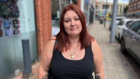 A woman with red hair and a black vest stands on a street next to parked cars. Shop fronts are also visible on the left of the image.