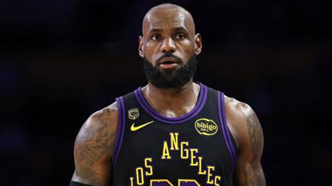 LeBron James stands in front of a black background. He is bald, has a black beard and has heavily tattooed arms. He is wearing a black basketball top which says Los Angeles in yellow letters.