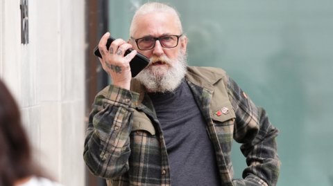 A man with long white hair, a full white beard and glasses holds a mobile phone to one ear while standing outside a building. The person is wearing a checked overshirt over a dark T-shirt, with tattoos visible on one hand.