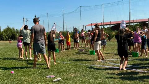 A group of people take part in an outdoor water game on a grassy field. Buckets, water balloons and plastic containers are scattered around, while participants stand in teams under a clear blue sky.