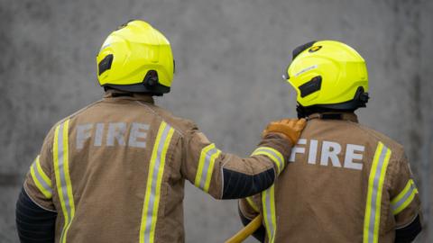 Two firefighters with their back to the camera. They are both wearing a high-vis yellow helmet and brown jackets with "fire" written on the back. One firefighter has their hand on the other's shoulder.