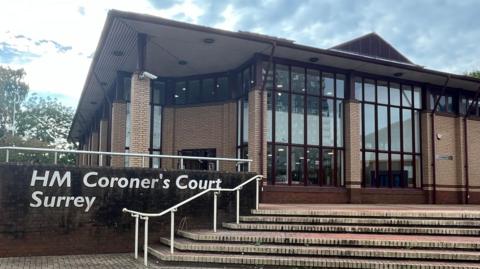 A general view of HM Coroner's Court Surrey. It is a brown brick building with outdoor stairs leading to the buildings facade which has long narrow windows. There are large white letters on a wall reading HM Coroner's Court Surrey.