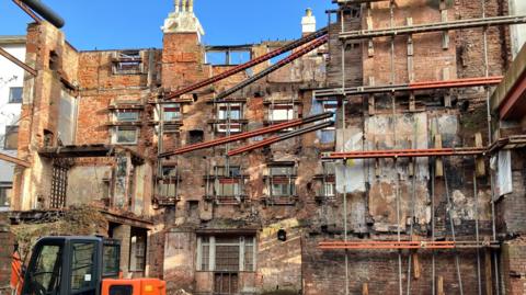 The remaining exterior walls and brickwork of an old fire ravaged building, with an orange and black digger in the foreground.