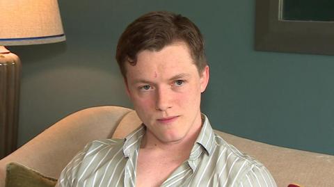 A man wearing a grey and white shirt sat on a cream sofa. There is a lamp in the background.
